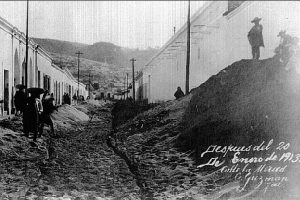 Town is covered by volcanic soot, with people investigating the piles of ash. A mountain can be seen in the backdrop.