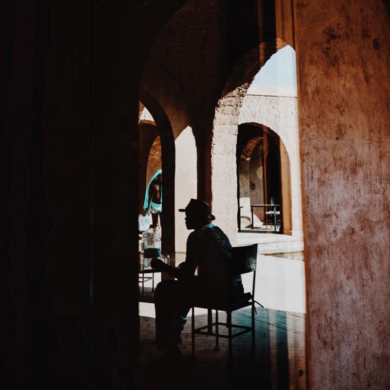 Man with fedora sits in the shadows and beige arches