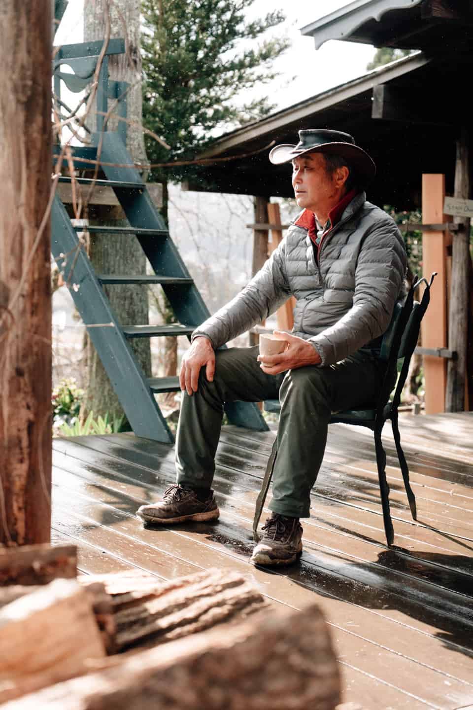 Elder man in cowboy hat sits on a chair with a coffee in hand as a sun shower comes down