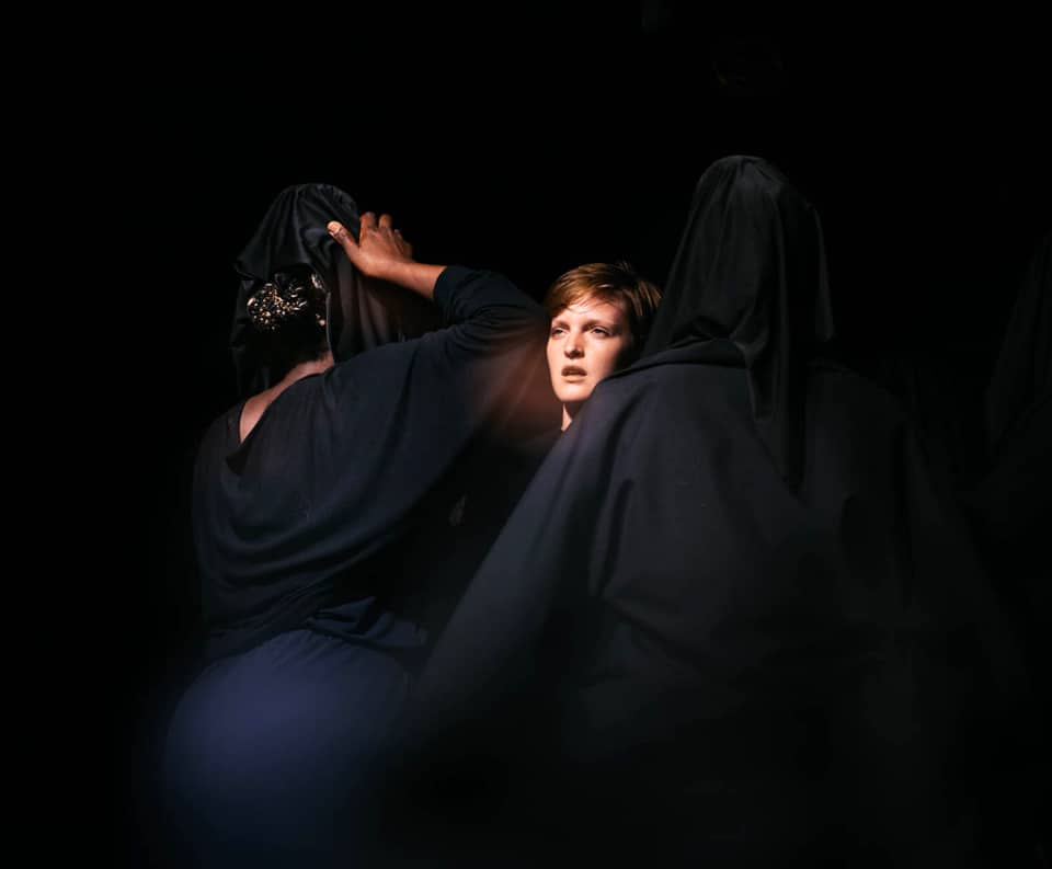 3 women huddled together one with their face visible the others shrouded in dark clothing