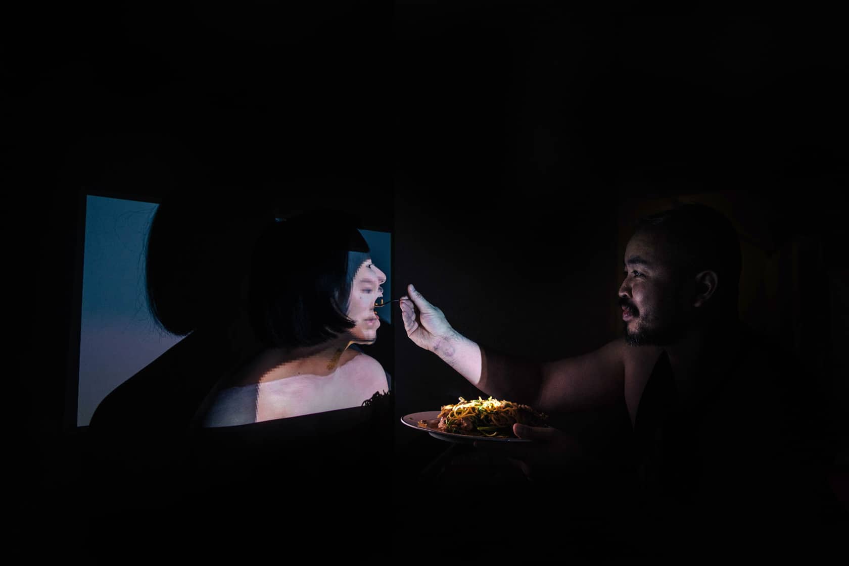 A person feed pasta to another person who is illuminated by a box